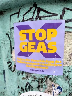 A sticker with the text 'STOP GEAS' in bold yellow letters on a purple background.  The smaller text below reads 'Gegen die Verschärfung des Gemeinsamen Europäischen Asylsystems!' which translates from German to "Against the tightening of the Common European Asylum System!"  A website address, stop-geas.de, is also included.