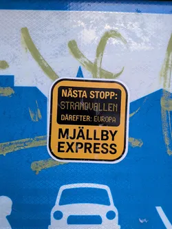 A rectangular sticker with rounded corners, featuring a bold yellow and black color scheme. The top and bottom sections have black text on a yellow background, while the middle section has yellow text on a black background. The text 'STRANDVALLEN' and 'EUROPA' appear in a pixelated, dot-matrix style font, mimicking an old-school departure board. The sticker has a thin white border around its edge and is applied to a textured, reflective blue and white surface with green graffiti.