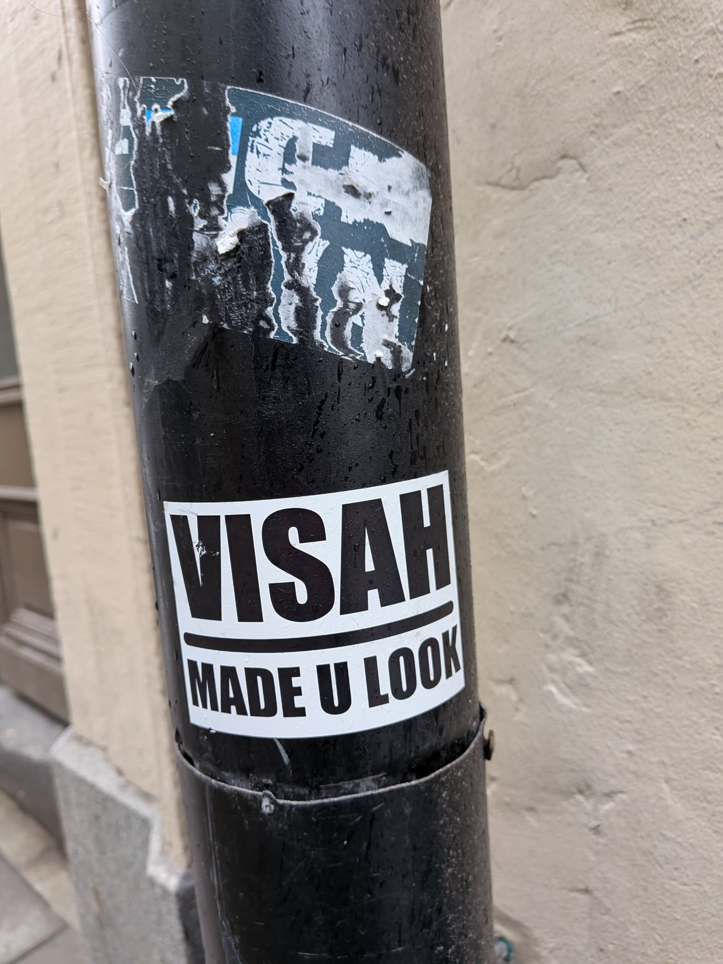 A rectangular, monochrome sticker featuring bold black text on a white background. The top line reads 'VISAH' in large, sans-serif capital letters. Below it, separated by a thin black line, the second line reads 'MADE U LOOK' in similar but slightly smaller capital letters. The sticker is applied to a dark, possibly black, metal drainpipe, showing some minor wear and tear, particularly around the edges and a small scuff on the letter 'S' in 'VISAH'.