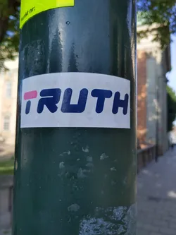 A sticker with the word \"TRUTH\" written in a stylized font. The letter \"I\" is pink, while the rest of the letters are navy blue. The sticker is attached to a dark green pole.