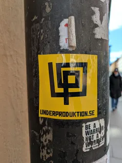 A sticker with a yellow background and a black logo consisting of interconnected squares and a stylized "LP" in the top portion. Below the logo is the text "UNDERPRODUKTION.SE". This is attached to a pole. Underneath, there is a second sticker which says "BE A WARRIOR NOT A WORRIER", followed by "www.realsoul.ca".