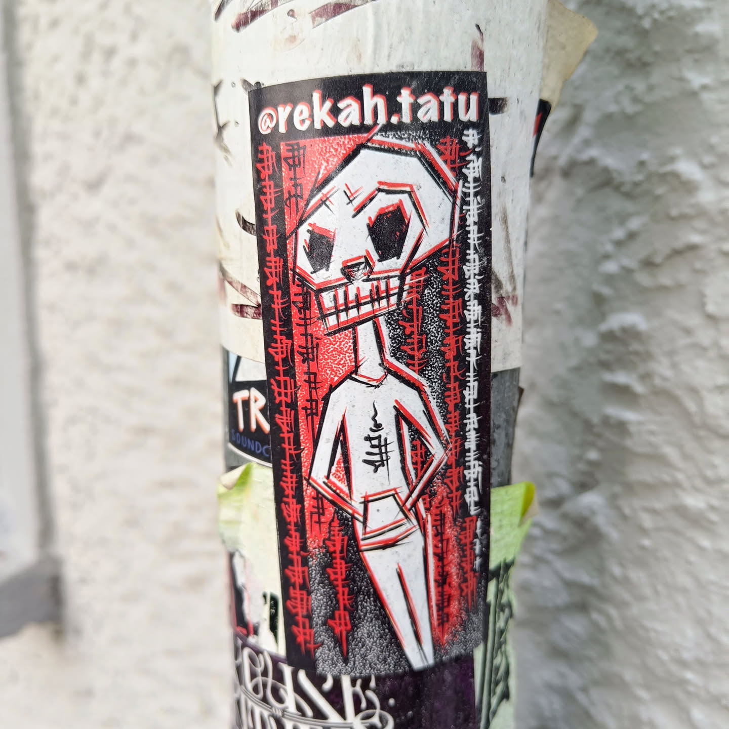 A sticker featuring a stylized skull character in red and white. The character has a humanoid body and is depicted in a confident pose.  The sticker has a black border with the artist's Instagram handle, @rekah.tatu, at the top. There is text along the sides, possibly in Japanese or a similar East Asian script. The art style is bold and graphic.