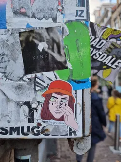 Sticker of a girl wearing a red bucket hat, with her hand covering one eye, seemingly crying. The text 'la joissance' is written underneath her chin.  The sticker is affixed to a weathered surface covered in other stickers and graffiti.