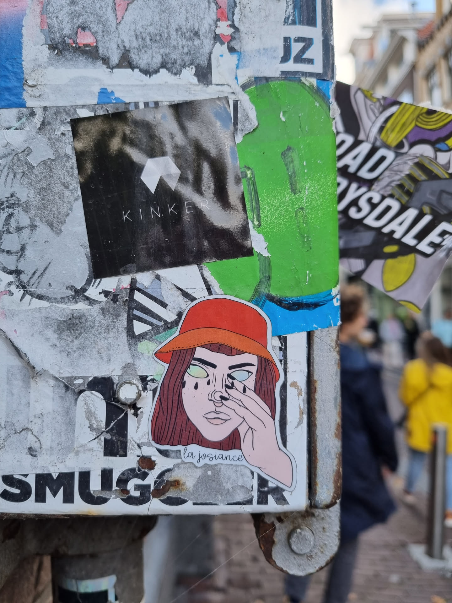 Sticker of a girl wearing a red bucket hat, with her hand covering one eye, seemingly crying. The text 'la joissance' is written underneath her chin.  The sticker is affixed to a weathered surface covered in other stickers and graffiti.