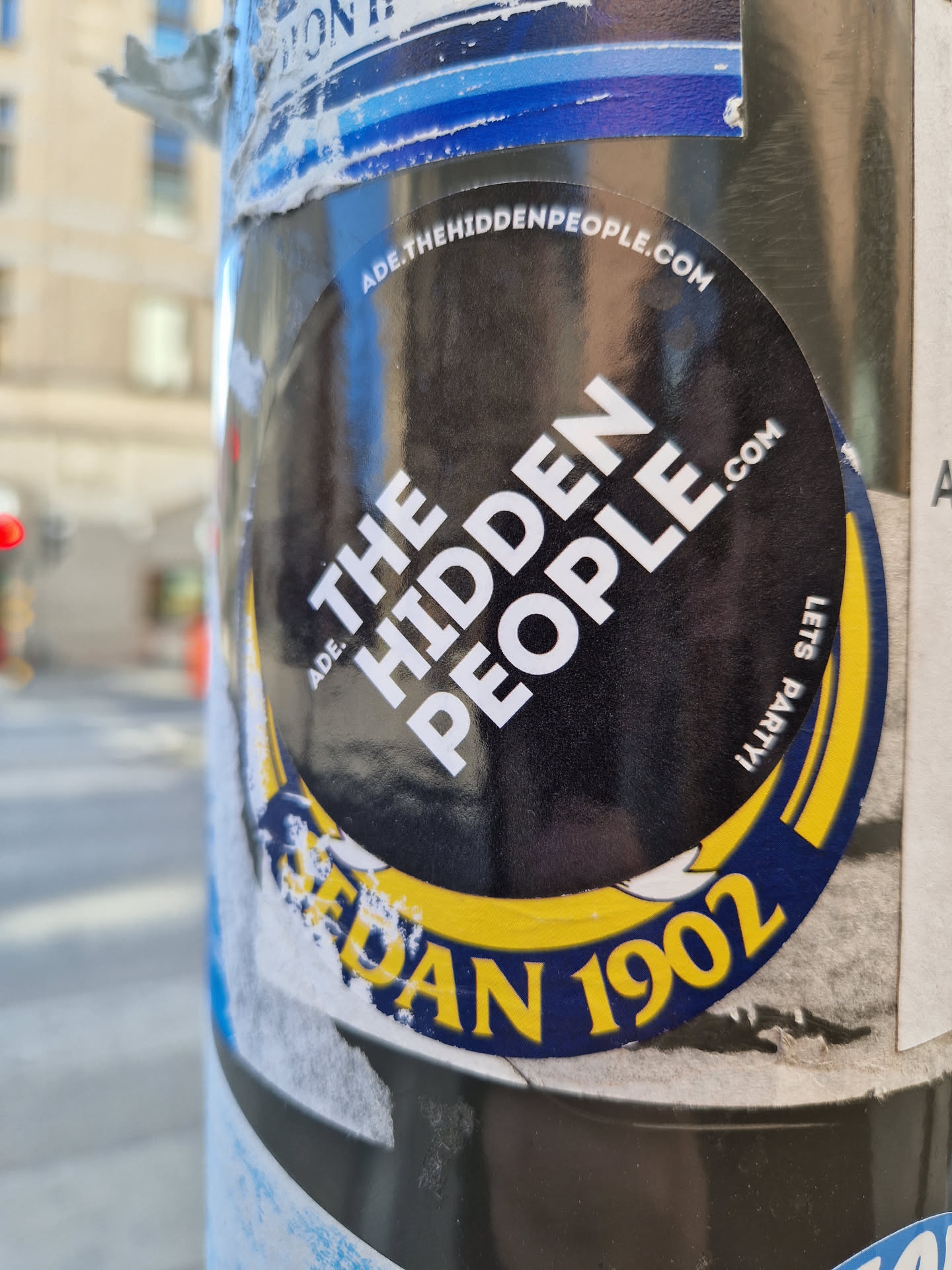 Round sticker with the text "The Hidden People" in the center.  The sticker has a yellow and blue border with the text "ADE. SEDAN 1902" visible at the bottom.  A website address is visible around the top edge: ADE.THEHIDDENPEOPLE.COM