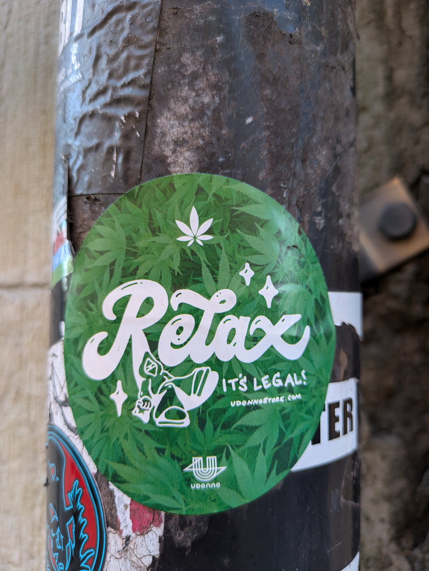 A round sticker with a green marijuana leaf background. It features white cursive text that says "Relax" and smaller text that says "It's Legal!" and a website address. There's also a small illustration of a person relaxing. The sticker has a logo with a stylized 'U'.