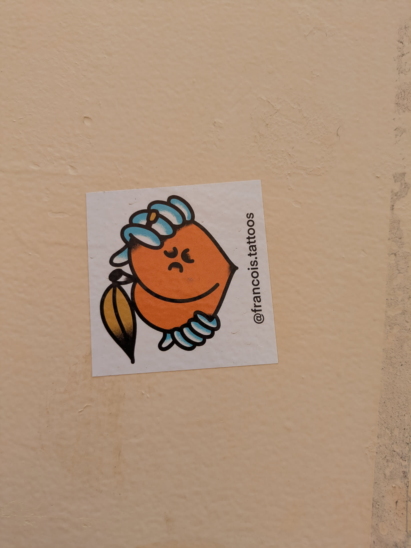 An orange, fruit-like character sticker with a leaf is placed on a wall. The character has a gloomy expression with downturned eyebrows and mouth, as well as hands in the form of semicircles. A leaf is seen on the left side.