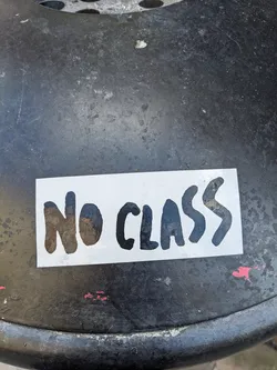 A white sticker with the text "NO CLASS" in black, stylized lettering. The sticker is affixed to a dark gray or black surface, possibly metal.
