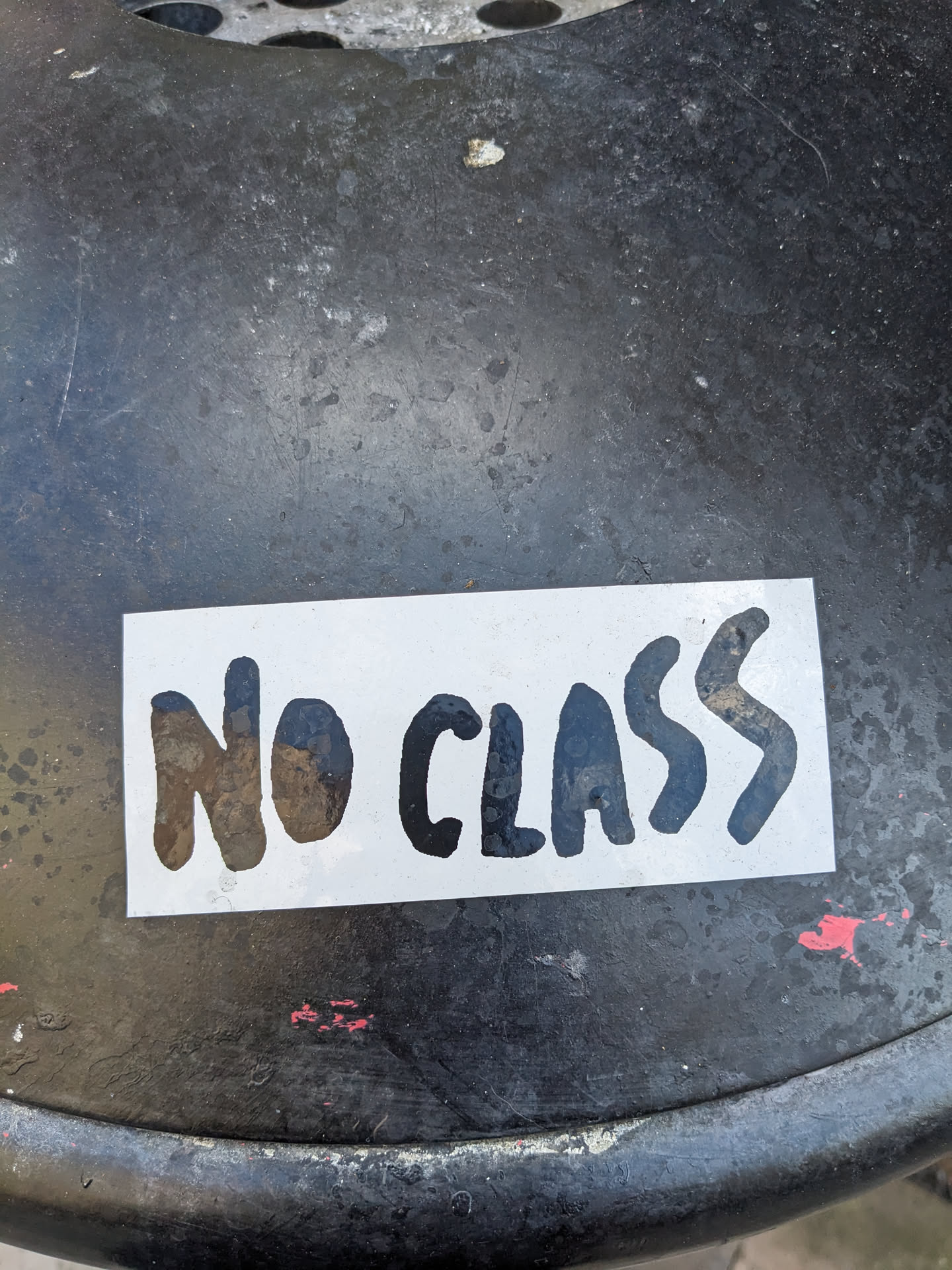 A white sticker with the text "NO CLASS" in black, stylized lettering. The sticker is affixed to a dark gray or black surface, possibly metal.