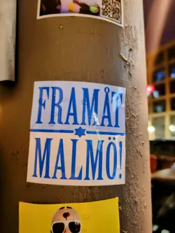 A sticker with the text "Framåt Malmö!" (Forward Malmö!) in blue letters on a white background. There's a small star of David symbol above the text.  The sticker is affixed to a weathered pole.