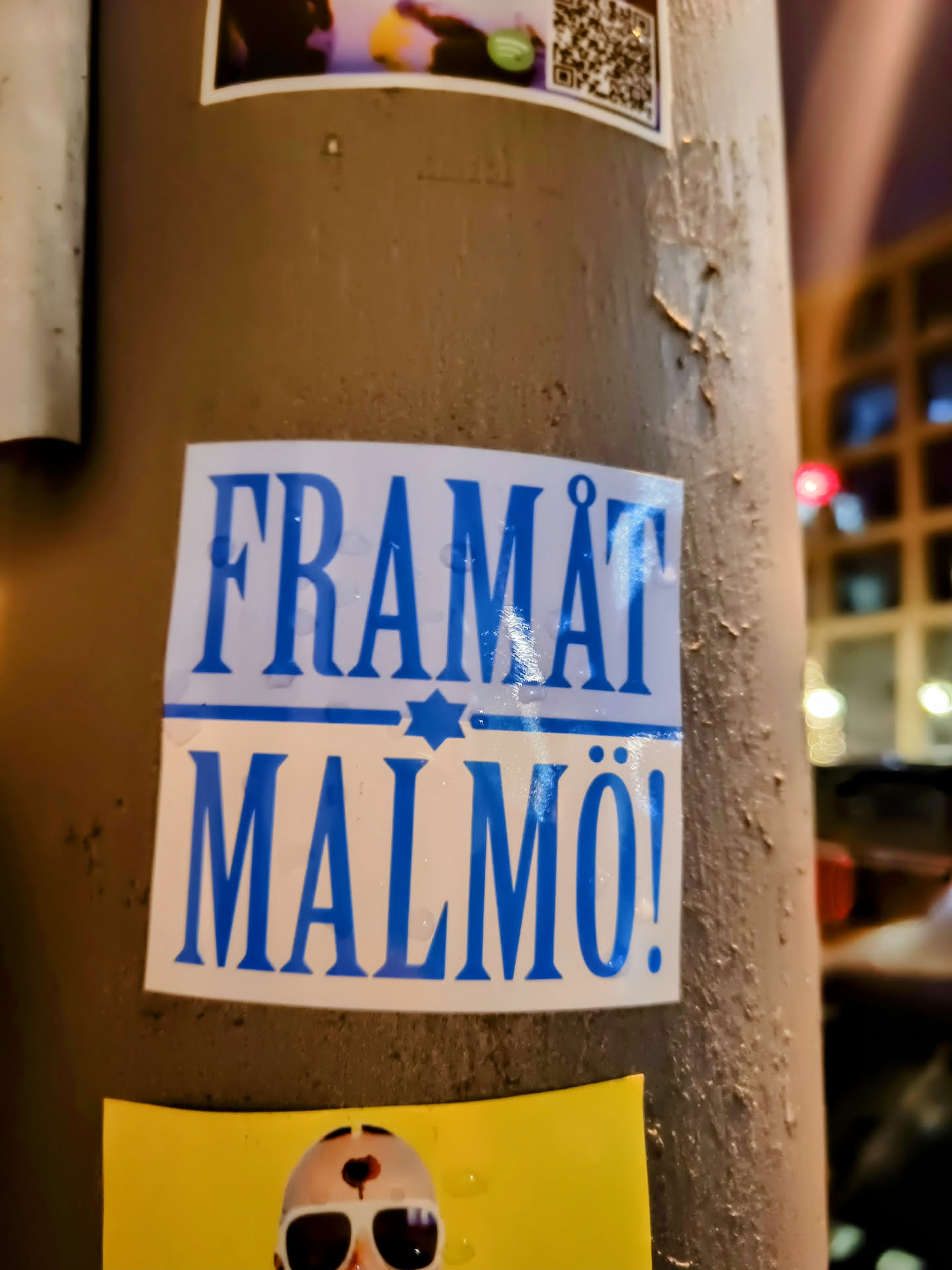 A sticker with the text "Framåt Malmö!" (Forward Malmö!) in blue letters on a white background. There's a small star of David symbol above the text.  The sticker is affixed to a weathered pole.
