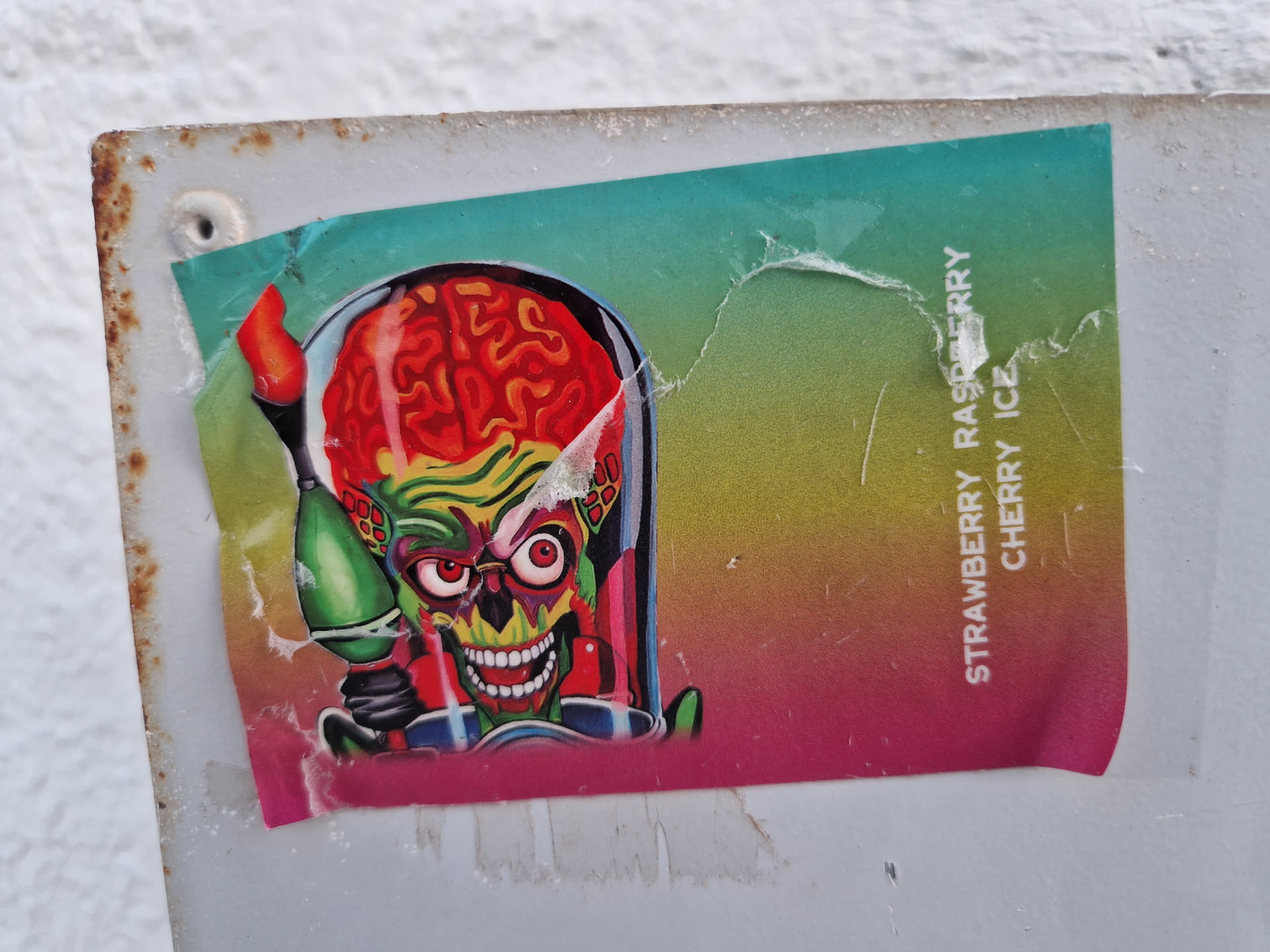 A sticker featuring the iconic Mars Attacks alien with a ray gun. The sticker has a colorful, gradient background with text indicating the flavors "Strawberry Raspberry Cherry Ice". The sticker shows some wear and tear from being affixed to a surface.