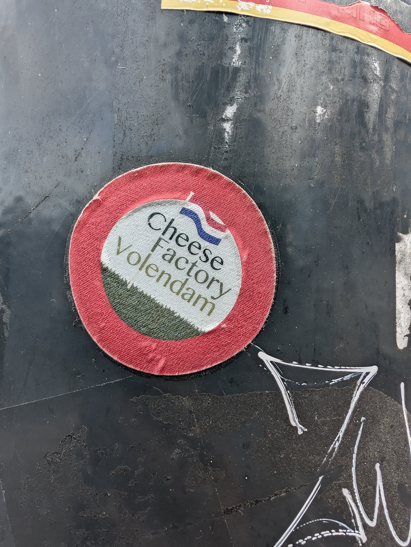 A round sticker with a red border and a white and green center. The text on the sticker says "Cheese Factory Volendam" and includes a small Dutch flag. The sticker is affixed to a dark gray surface.