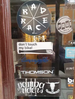 A collection of stickers on a glass door.  The main focus is a Pampa Riders Cycling Club sticker featuring a skull logo. Other stickers include one that says "don't touch my bike!" and several bike part brand logos.