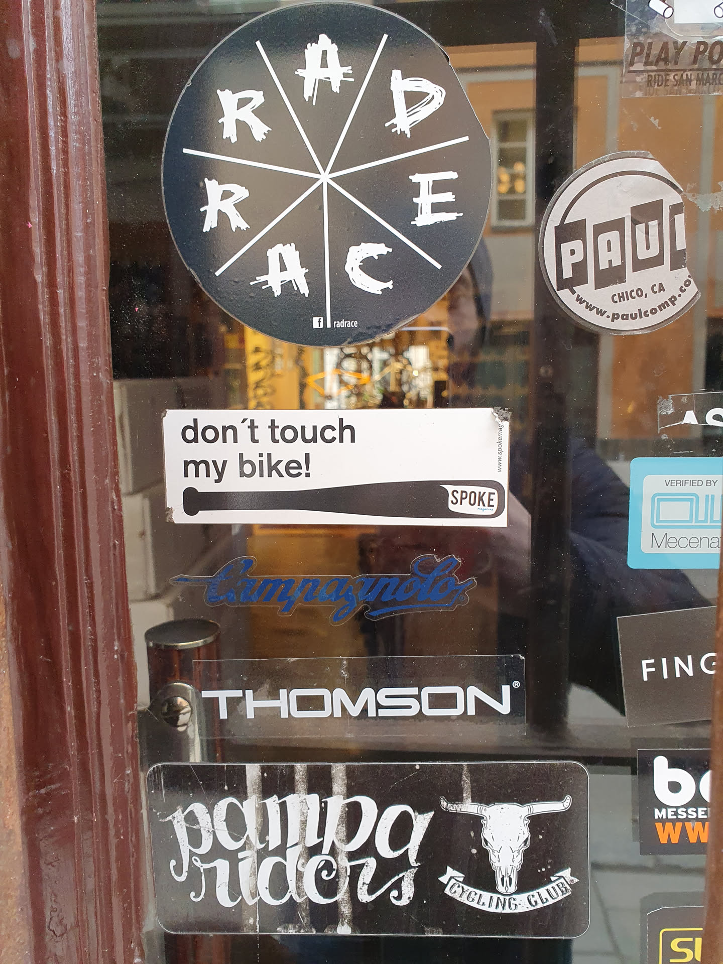 A collection of stickers on a glass door.  The main focus is a Pampa Riders Cycling Club sticker featuring a skull logo. Other stickers include one that says "don't touch my bike!" and several bike part brand logos.