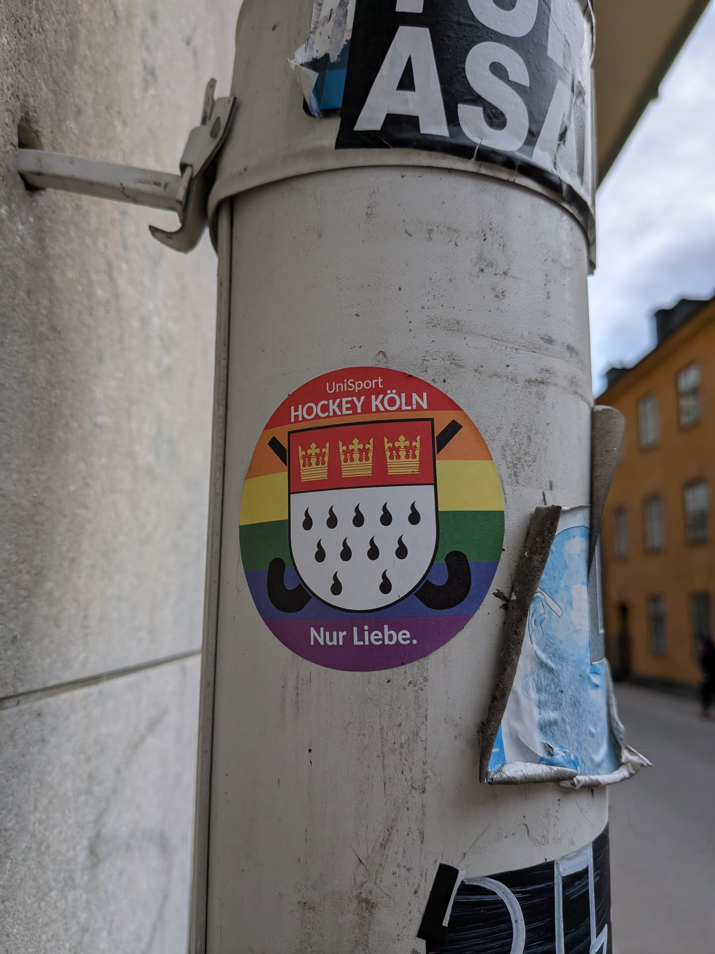 A circular sticker featuring a vibrant rainbow background with horizontal stripes. In the center, a shield-like emblem is depicted, with its upper half red containing three golden crowns, and its lower half white with eleven black teardrop shapes. Behind the shield, two black hockey sticks are crossed. Arched above the emblem on a red band is the white text 'UniSport HOCKEY KÖLN', and below on a purple band is the white text 'Nur Liebe.'. The sticker is applied to a light-colored pole, showing slight wear and tear.
