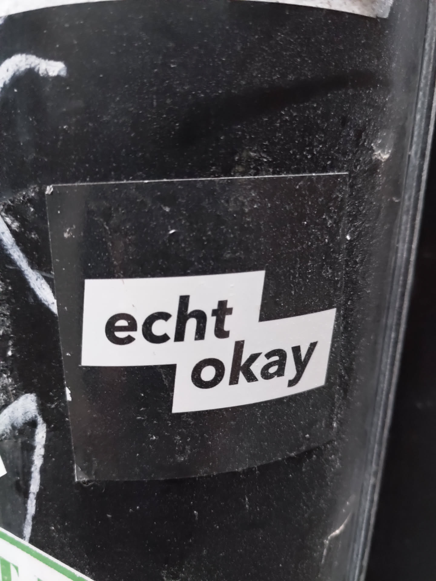 A black and white sticker with the text "echt okay" on it. The text is split into two lines, with "echt" on the top line and "okay" on the bottom line. The sticker is attached to a dark-colored surface.