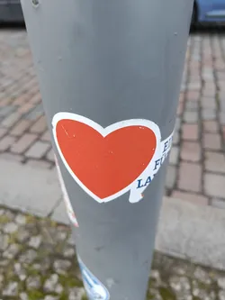 A simple yet effective sticker with a red heart shape on a white background. The sticker is placed on a grey pole outdoors. Part of another sticker is visible next to the red heart.