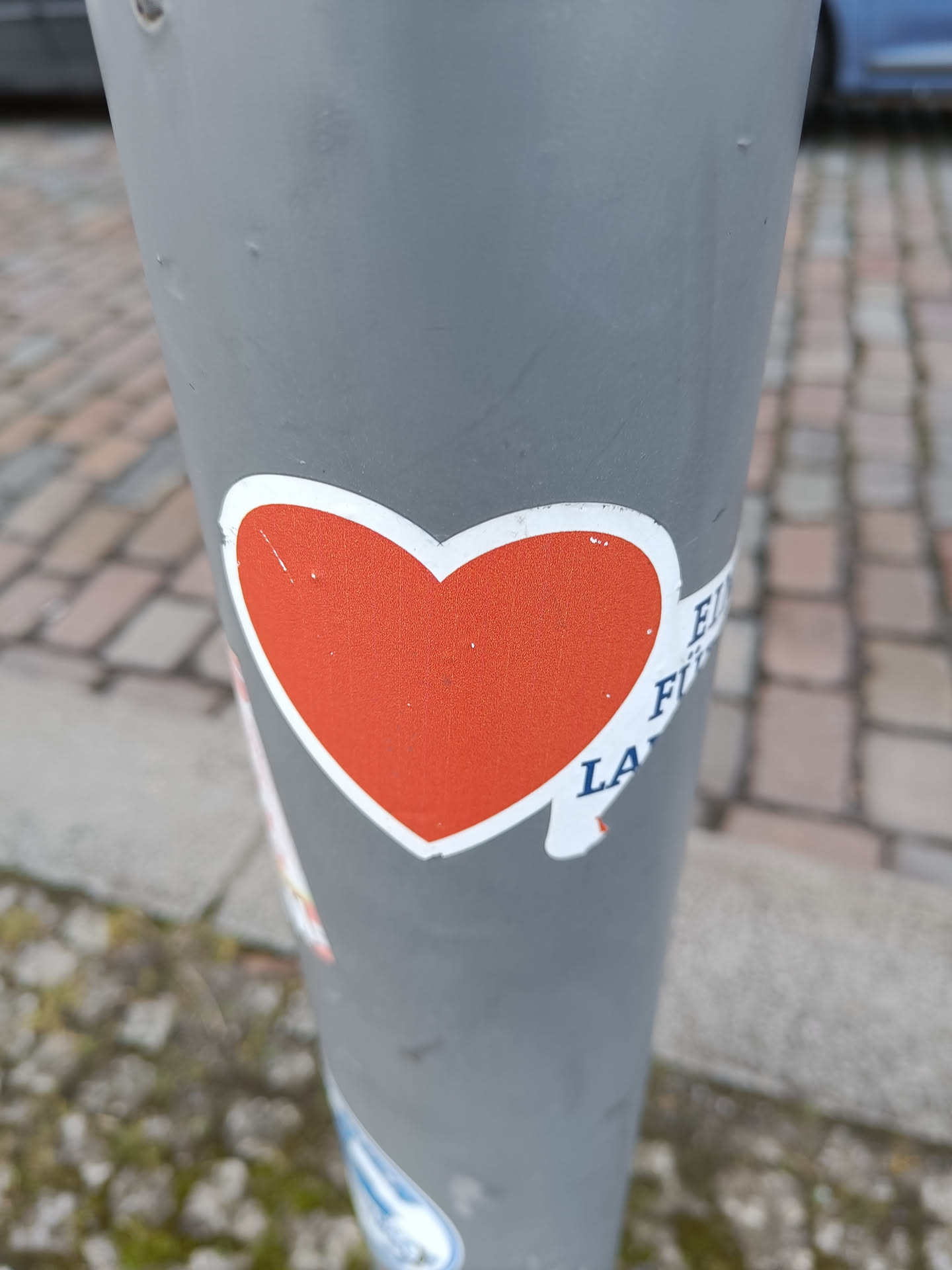 A simple yet effective sticker with a red heart shape on a white background. The sticker is placed on a grey pole outdoors. Part of another sticker is visible next to the red heart.
