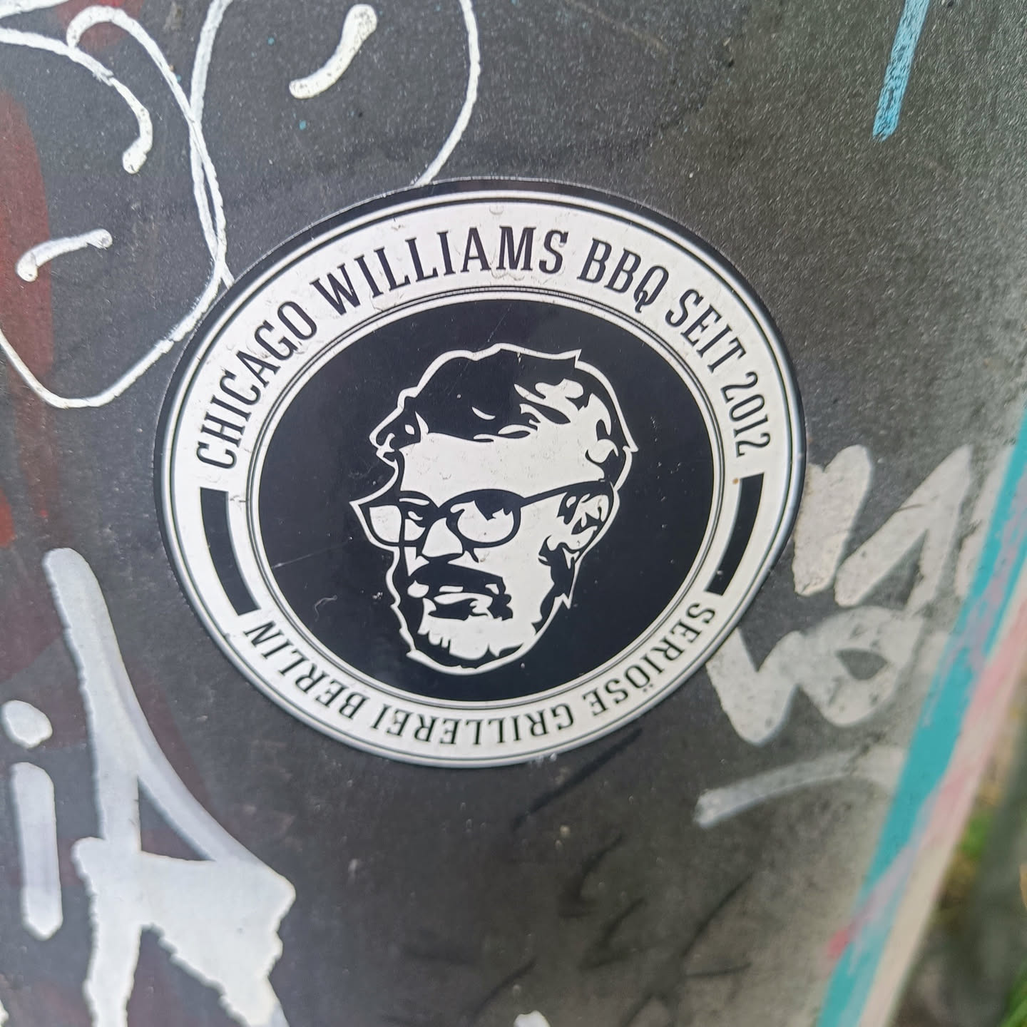 A round black and white sticker with the text "Chicago Williams BBQ Seit 2012" and "Seriöse Grillerei Berlin" in a circular pattern.  In the center is a black and white illustration of a man with glasses and a beard. 