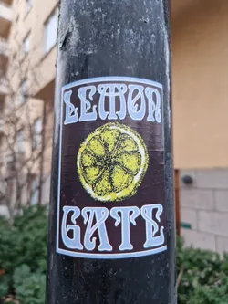 A sticker featuring a lemon and the text "Lemon Gate" in a retro style font. The sticker is on a dark brown background with a light blue border.