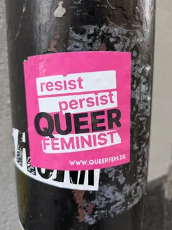 Pink sticker with the text "resist persist QUEER FEMINIST" and the website www.queerfem.de. It's attached to a dark pole.