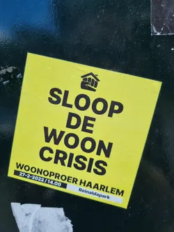 A bright yellow sticker advocating for the demolition of the housing crisis.  It features a fist in a house logo and the date and location of a protest in Haarlem, Netherlands. 