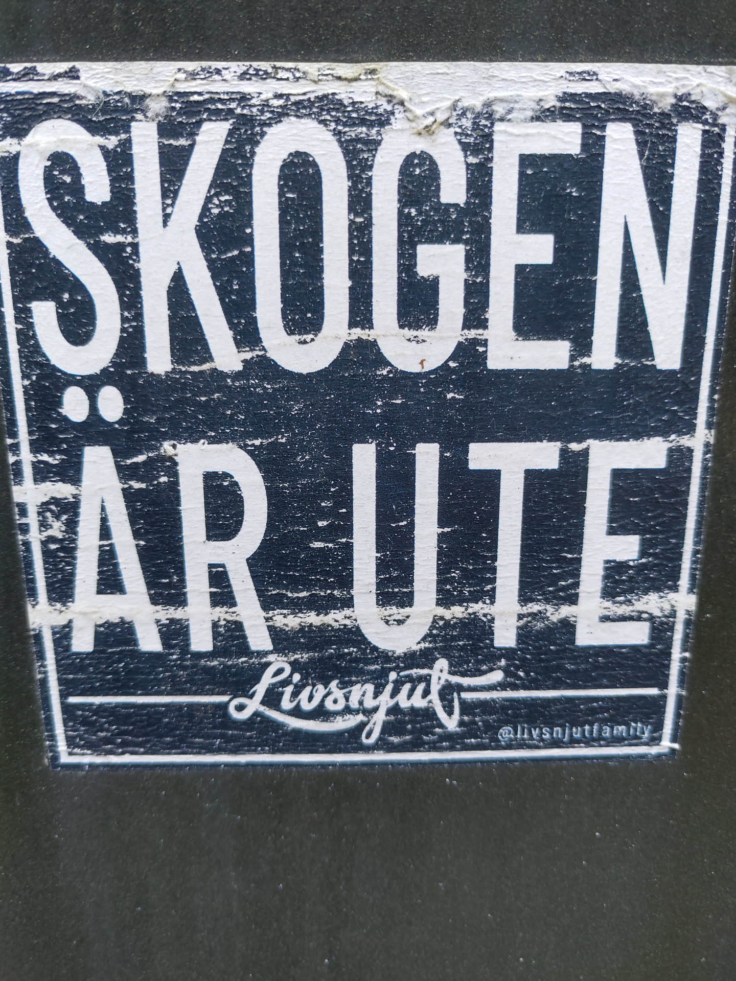 A dark blue sticker with white text. The text says "SKOGEN ÄR UTE" (The forest is out) and has the signature "Livsnjut" with the Instagram handle @livsnjutfamily. The sticker shows some signs of wear.