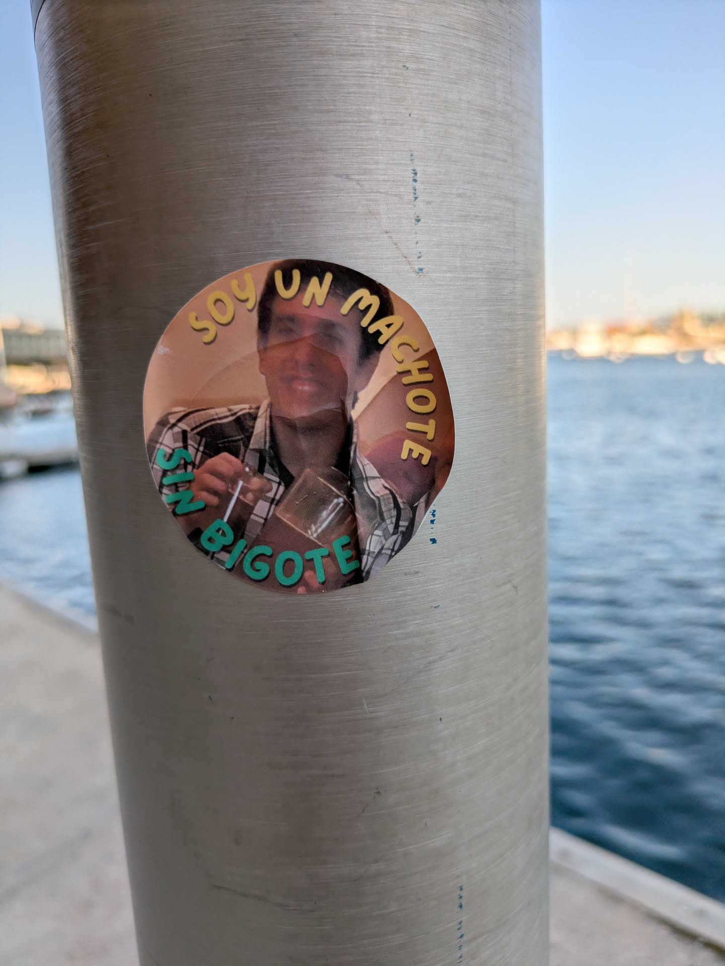 A round sticker adhered to a gray pole near the water, displaying a man holding a drink. The text 'Soy un machote sin bigote' is written around the image, creating a humorous effect.