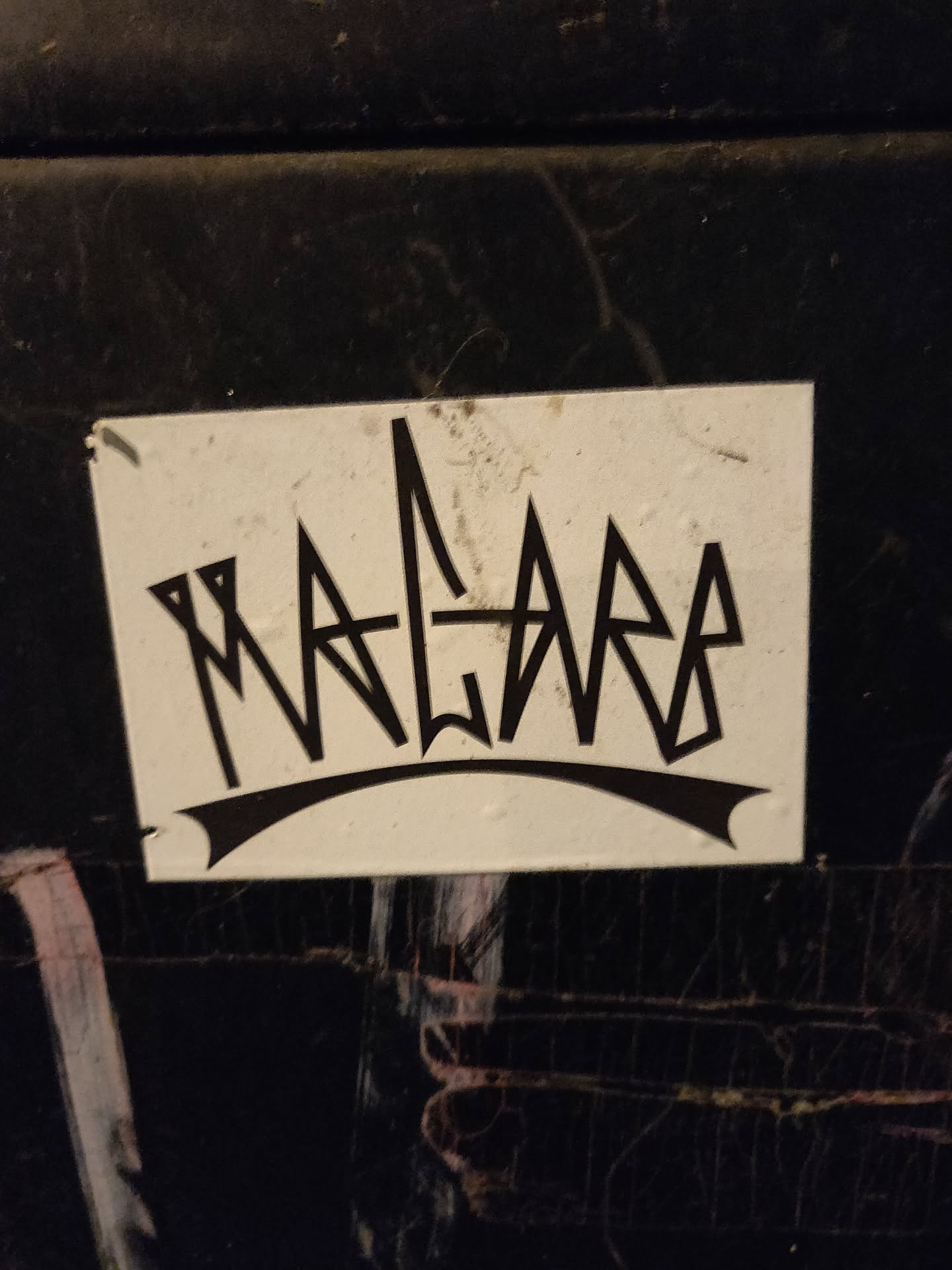 A white square sticker with black lettering. The word 'MALARA' is written in a stylized, almost graffiti-like font. The letters are sharp and angular, with a crown-like shape above them.  The sticker appears to be adhered to a dark surface.
