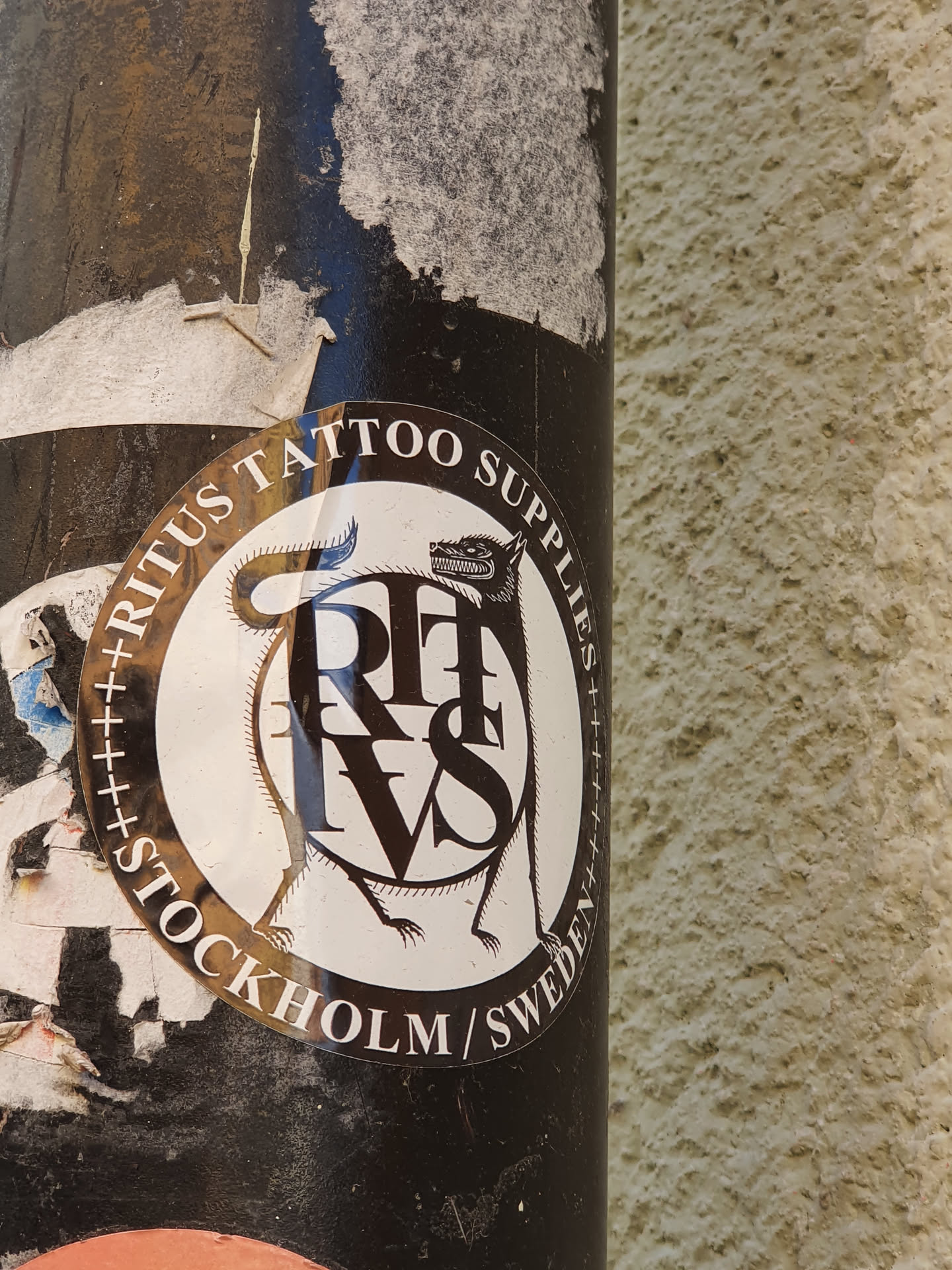 A round sticker with a black and white illustration of a wolf-like creature encircling the letters RITVS. The text "Ritvs Tattoo Supplies Stockholm/Sweden" is written around the circular design. The sticker appears to be affixed to a dark-colored pole.