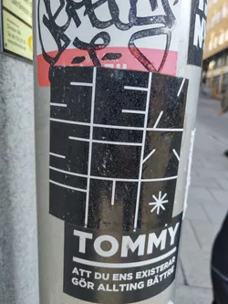 A black and white sticker with a grid-like pattern and the word "TOMMY" at the bottom. Below the brand name, there is Swedish text that reads "ATT DU ENS EXISTERAR GÖR ALLTING BÄTTRE", which translates to "THAT YOU EVEN EXIST MAKES EVERYTHING BETTER".