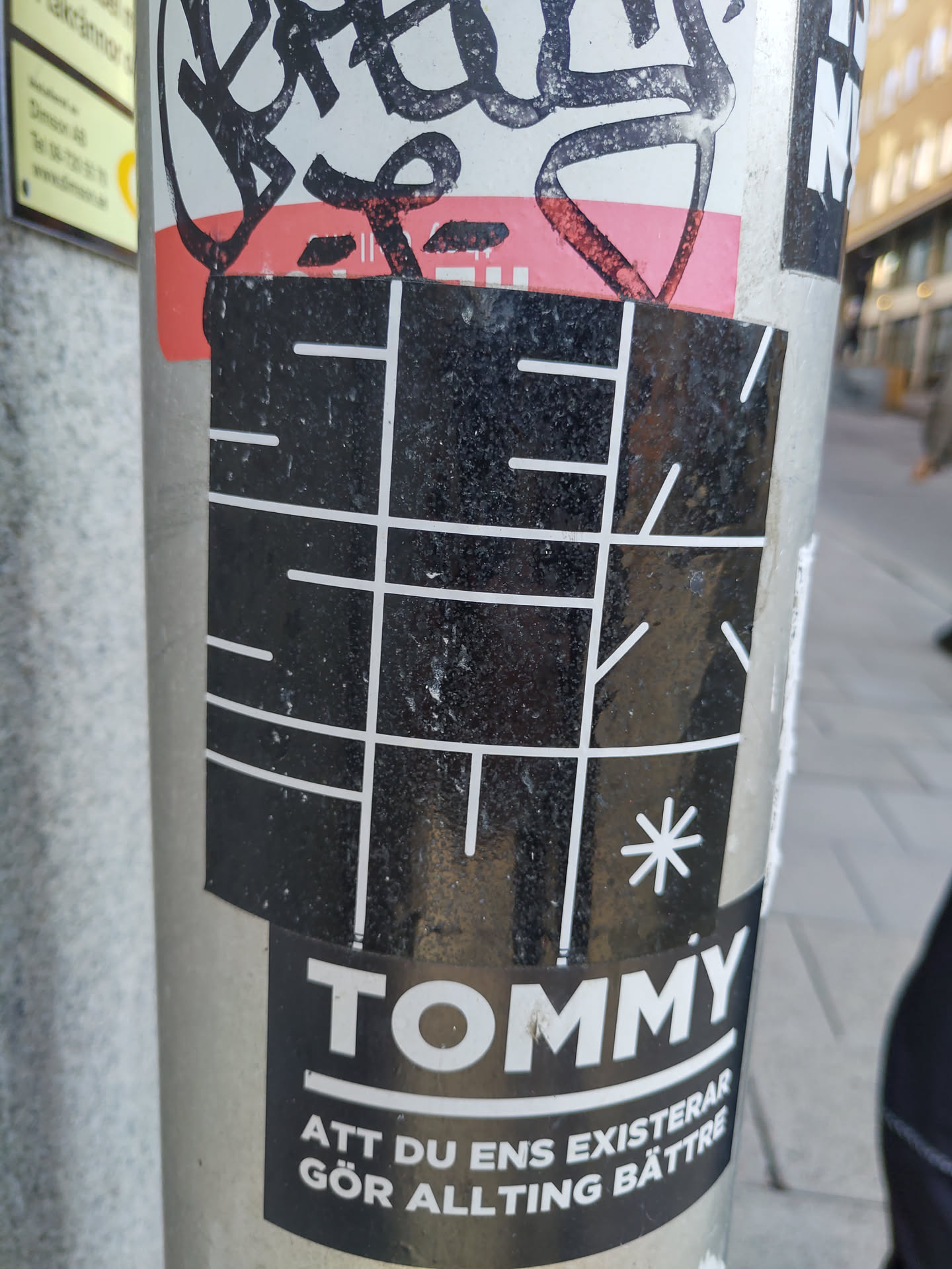 A black and white sticker with a grid-like pattern and the word "TOMMY" at the bottom. Below the brand name, there is Swedish text that reads "ATT DU ENS EXISTERAR GÖR ALLTING BÄTTRE", which translates to "THAT YOU EVEN EXIST MAKES EVERYTHING BETTER".