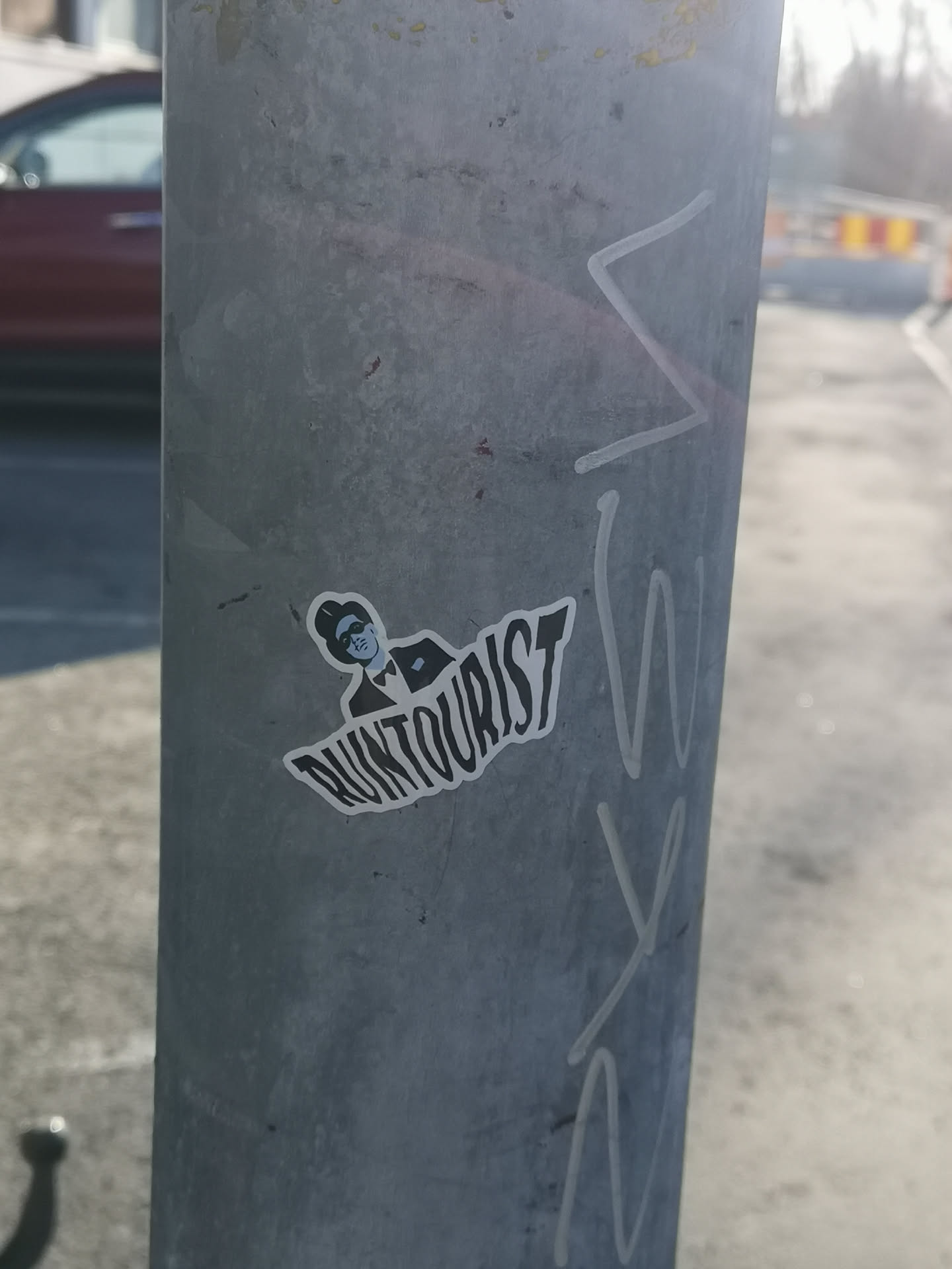 The sticker features a stylized, monochromatic illustration of a person, likely male, from the chest up. The figure wears a dark fedora-style hat, dark sunglasses, a dark suit jacket, and a dark bow tie. The face is depicted with minimal features, primarily the sunglasses and a defined jawline, in a lighter blue-grey tone. The entire illustration is outlined in white, giving it a distinct cut-out appearance. Below the figure, the word "RUNTourist" is written in a bold, somewhat distressed or wavy sans-serif font, also in dark grey/black with a white outline. The overall aesthetic is clean yet edgy, with a limited color palette of black, white, and a muted blue-grey.