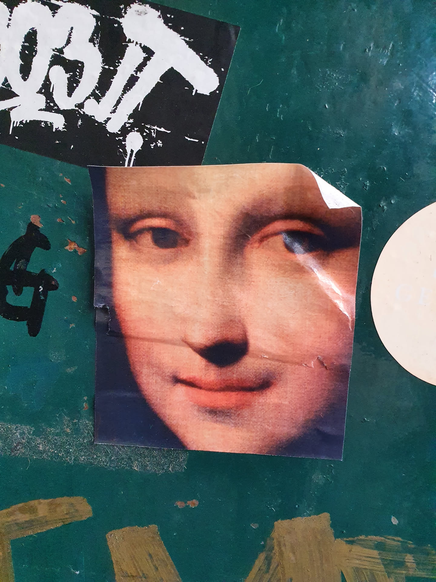 A sticker featuring a close-up of the Mona Lisa's face. The sticker has a slightly worn look, suggesting it may have been placed outdoors for some time. The background is a dark teal surface with other graffiti and stickers visible.