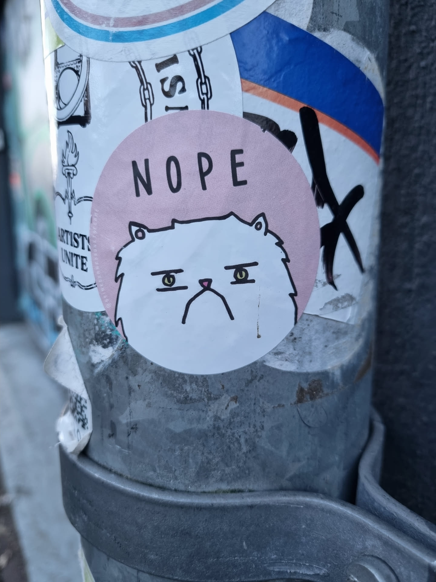 A round sticker featuring a grumpy-looking white cat with the word "NOPE" written in black sans-serif lettering on a pale pink background. The sticker is affixed to a metal pole, which also has other stickers on it.