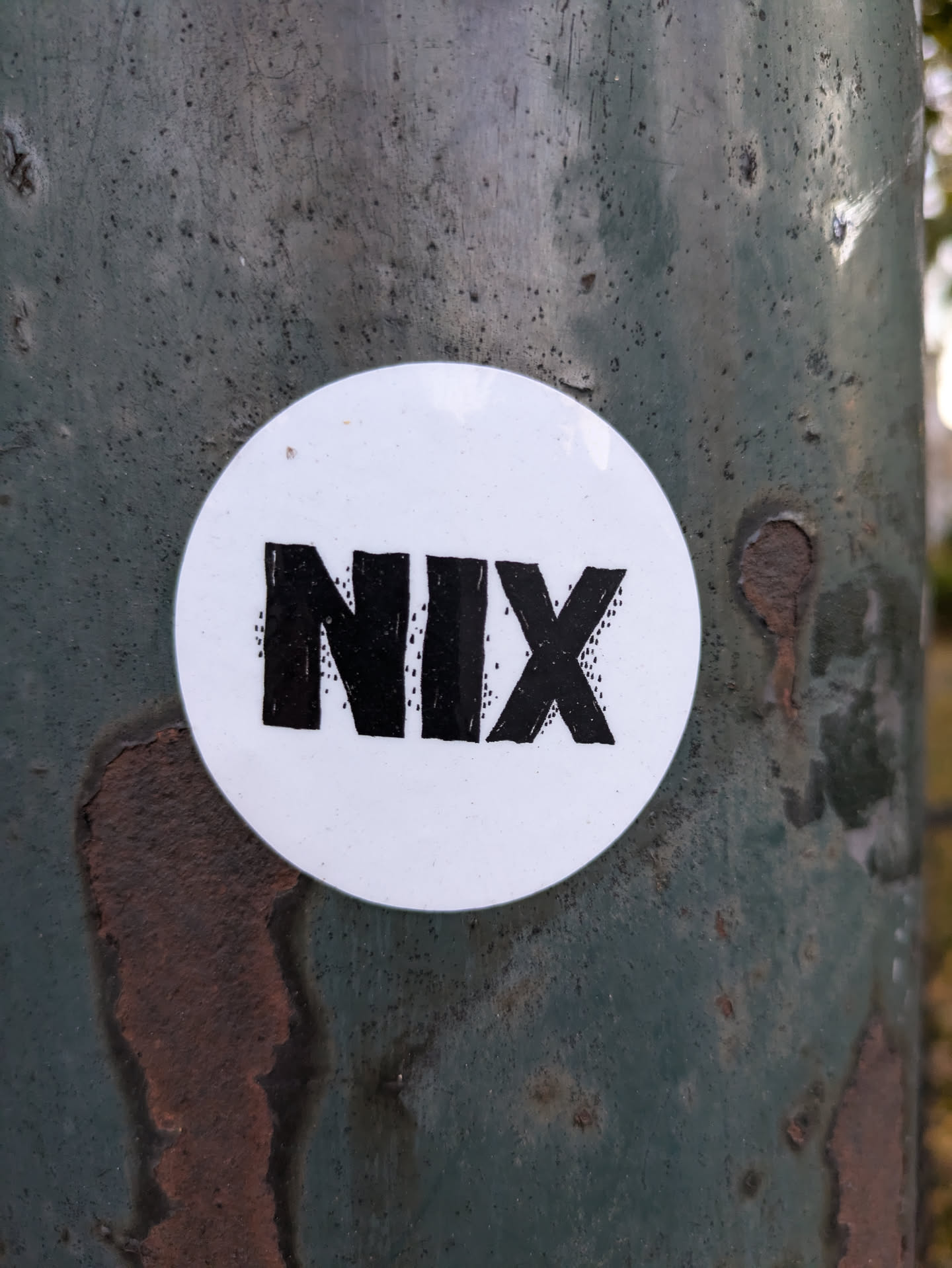 A round white sticker with the word NIX written in bold, black, stylized lettering. The sticker is affixed to a weathered, dark teal metal pole.