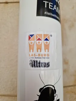 A white sticker with three stylized figures and the text "LAS-BURG Liefer-Adress-Service Ultras". The figures have colored triangles on top.