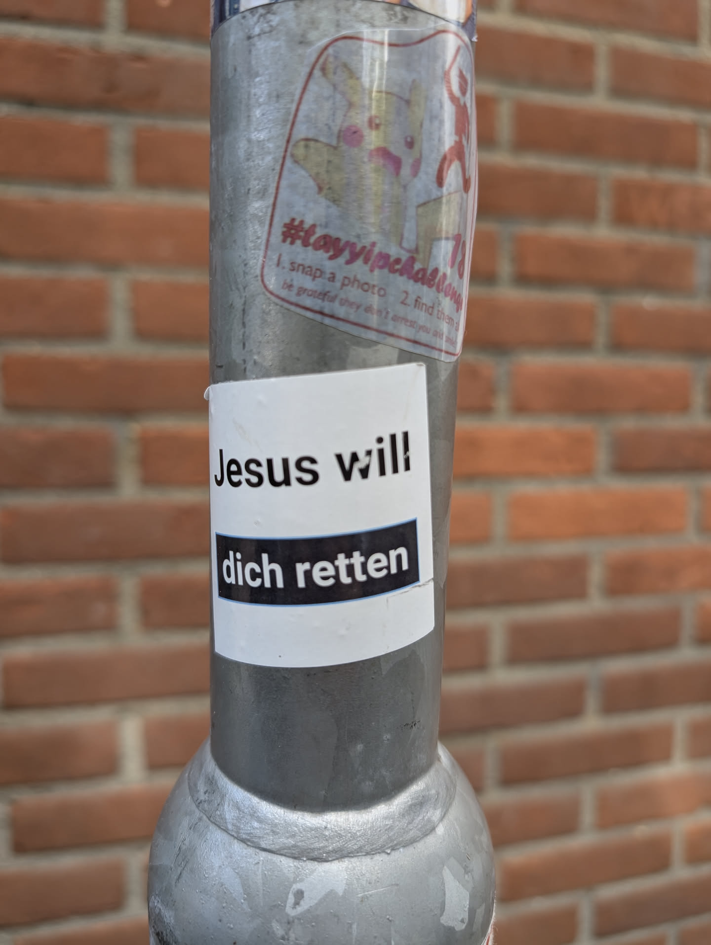 A white sticker with black text affixed to a metal pole. The text reads "Jesus will dich retten". The phrase translates to "Jesus will save you". There is another sticker above it. It appears to be a Pikachu character.