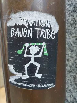 Black and white sticker with a drawing of a person holding a green and white scarf. The text 'International Bajen Tribe' is written at the top. At the bottom, there is some smaller text that seems to be locations: "SODER - ORTEN - ARSTA - GULLMARSPLAN".