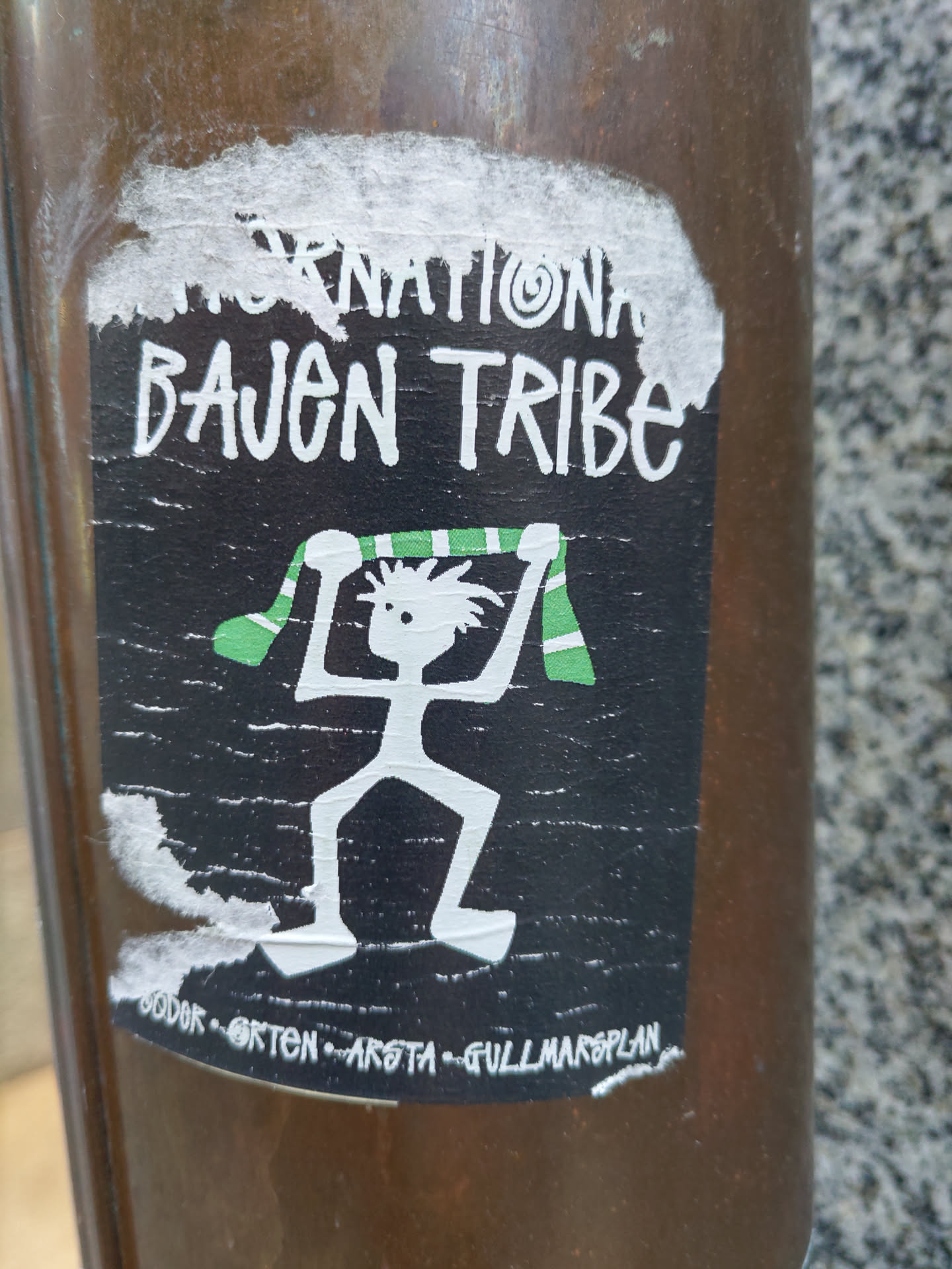 Black and white sticker with a drawing of a person holding a green and white scarf. The text 'International Bajen Tribe' is written at the top. At the bottom, there is some smaller text that seems to be locations: "SODER - ORTEN - ARSTA - GULLMARSPLAN".