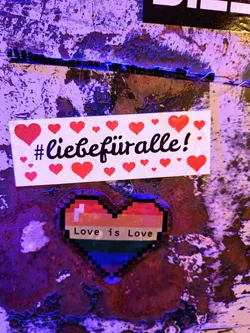 A sticker with the text #liebefüralle (love for all) surrounded by red hearts and a pixelated rainbow heart with the text Love is Love. The sticker is placed on a grunge-like surface.