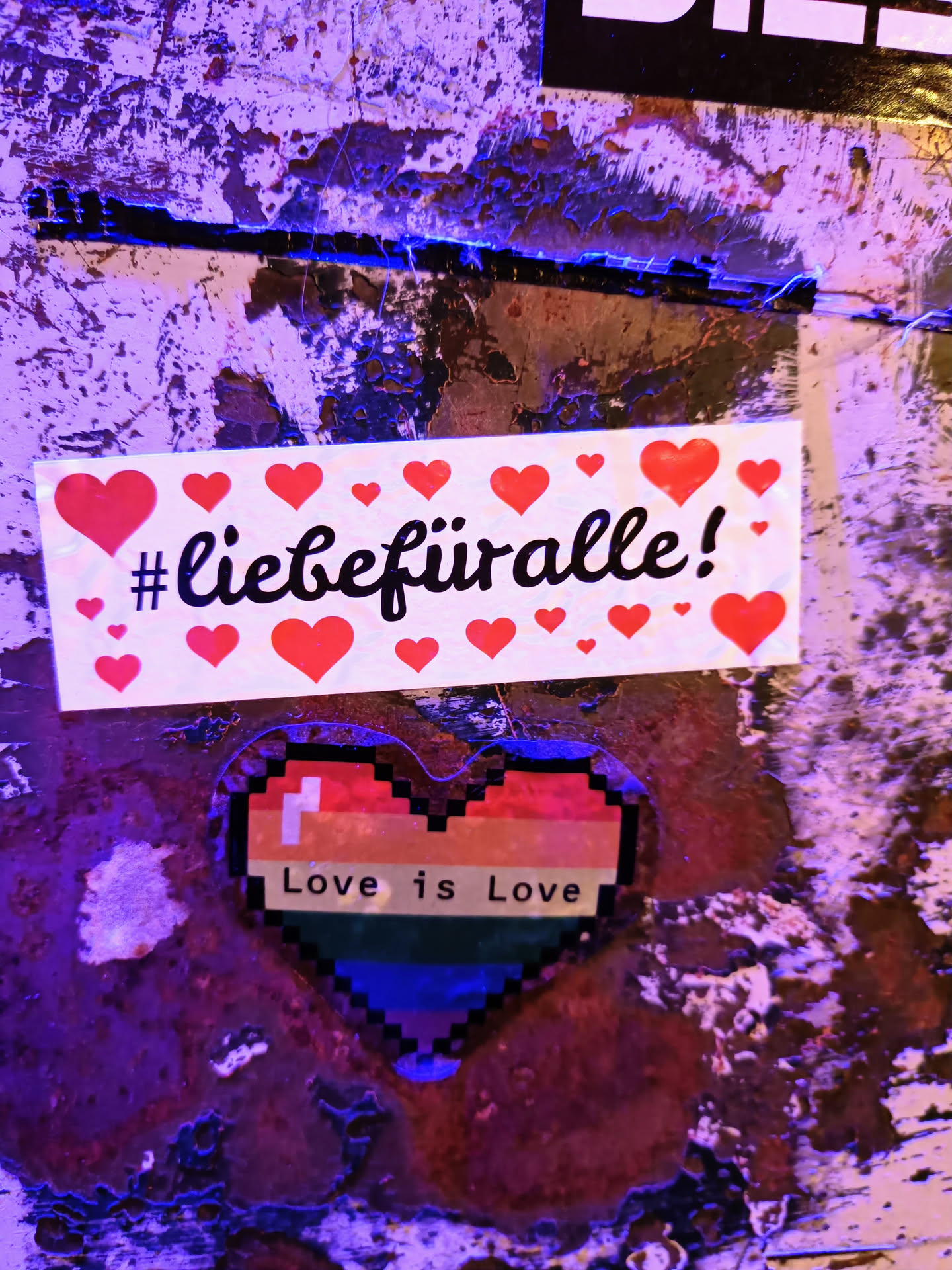 A sticker with the text #liebefüralle (love for all) surrounded by red hearts and a pixelated rainbow heart with the text Love is Love. The sticker is placed on a grunge-like surface.