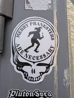 A die-cut black and white sticker formatted in the iconic silhouette of the Grateful Dead's 'Steal Your Face' skull. The circular top portion features an inner circle containing a black silhouette of a medieval court jester or fool running while holding a flower. Surrounding the jester along the circular border is the text 'MERRY PRANKSTERS' on top and 'ARE NECESSARY' along the bottom in a bold, stylized serif typeface. Below the circle is the black and white stylized jaw and teeth of the skull.