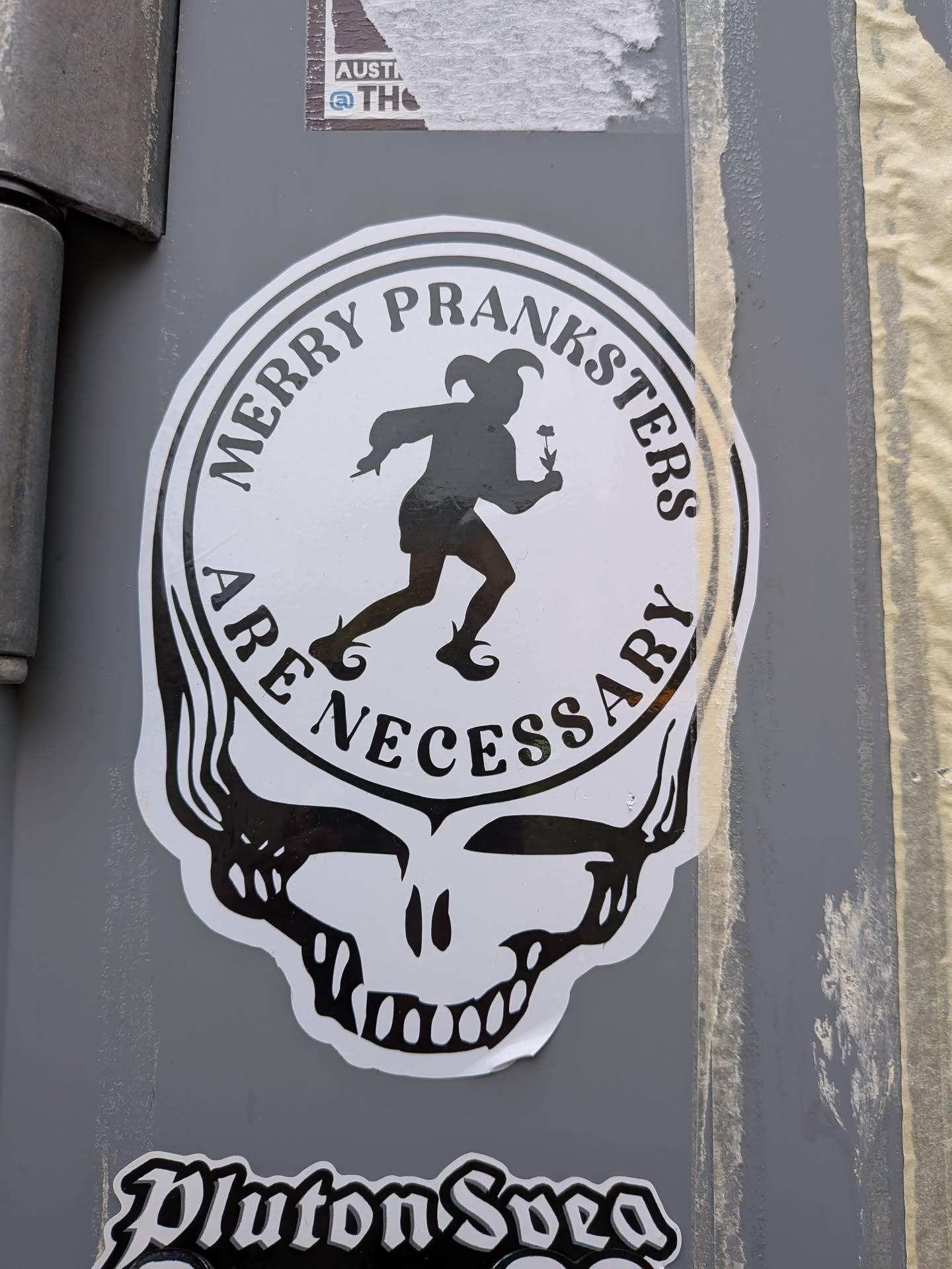 A die-cut black and white sticker formatted in the iconic silhouette of the Grateful Dead's 'Steal Your Face' skull. The circular top portion features an inner circle containing a black silhouette of a medieval court jester or fool running while holding a flower. Surrounding the jester along the circular border is the text 'MERRY PRANKSTERS' on top and 'ARE NECESSARY' along the bottom in a bold, stylized serif typeface. Below the circle is the black and white stylized jaw and teeth of the skull.