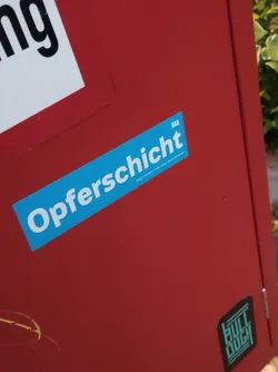 A light blue sticker with the word "Opferschicht" in white text on a red surface. There is a small logo in the top right corner of the sticker that says HAU. 