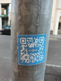A light blue square sticker with a QR code and the text "OnlyGrams" in the center is affixed to a metallic pole. The sticker appears to be promoting the OnlyGrams platform.