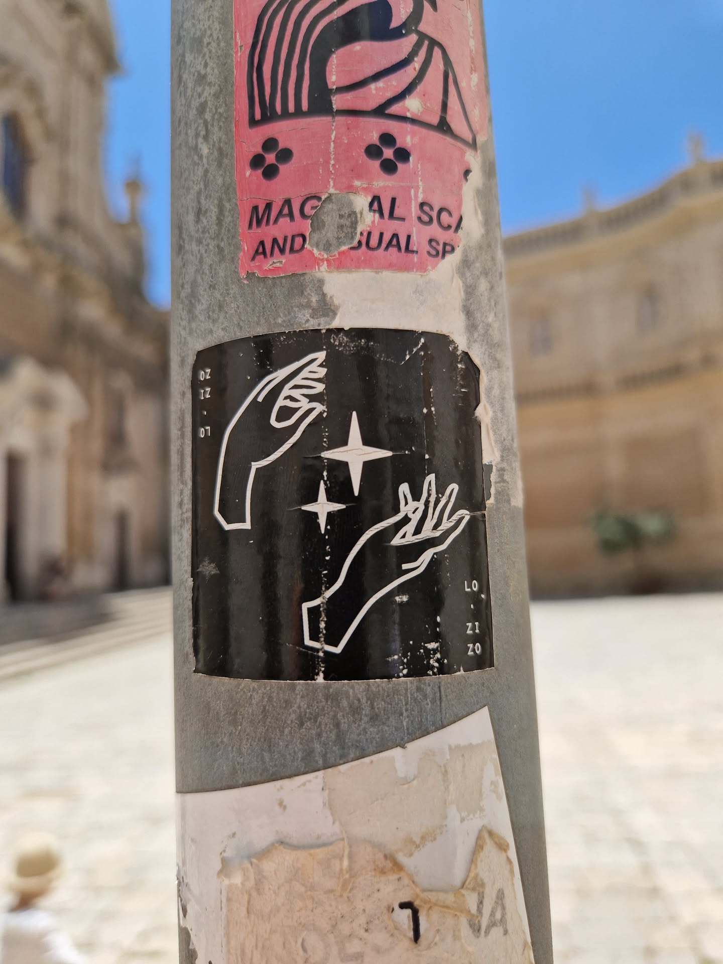 Black and white sticker with a minimalist design of two hands outstretched towards a shining star. The sticker is affixed to a metal pole, possibly outdoors. Some other damaged stickers are visible around it.