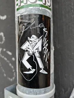 Black and white sticker featuring a stylized figure in a hat and loose-fitting clothing, possibly pants. The character's face is partially obscured, adding to the mysterious nature of the art. There are additional markings around the main figure, which could be considered part of the design or a signature. The sticker is affixed to a metallic pole.