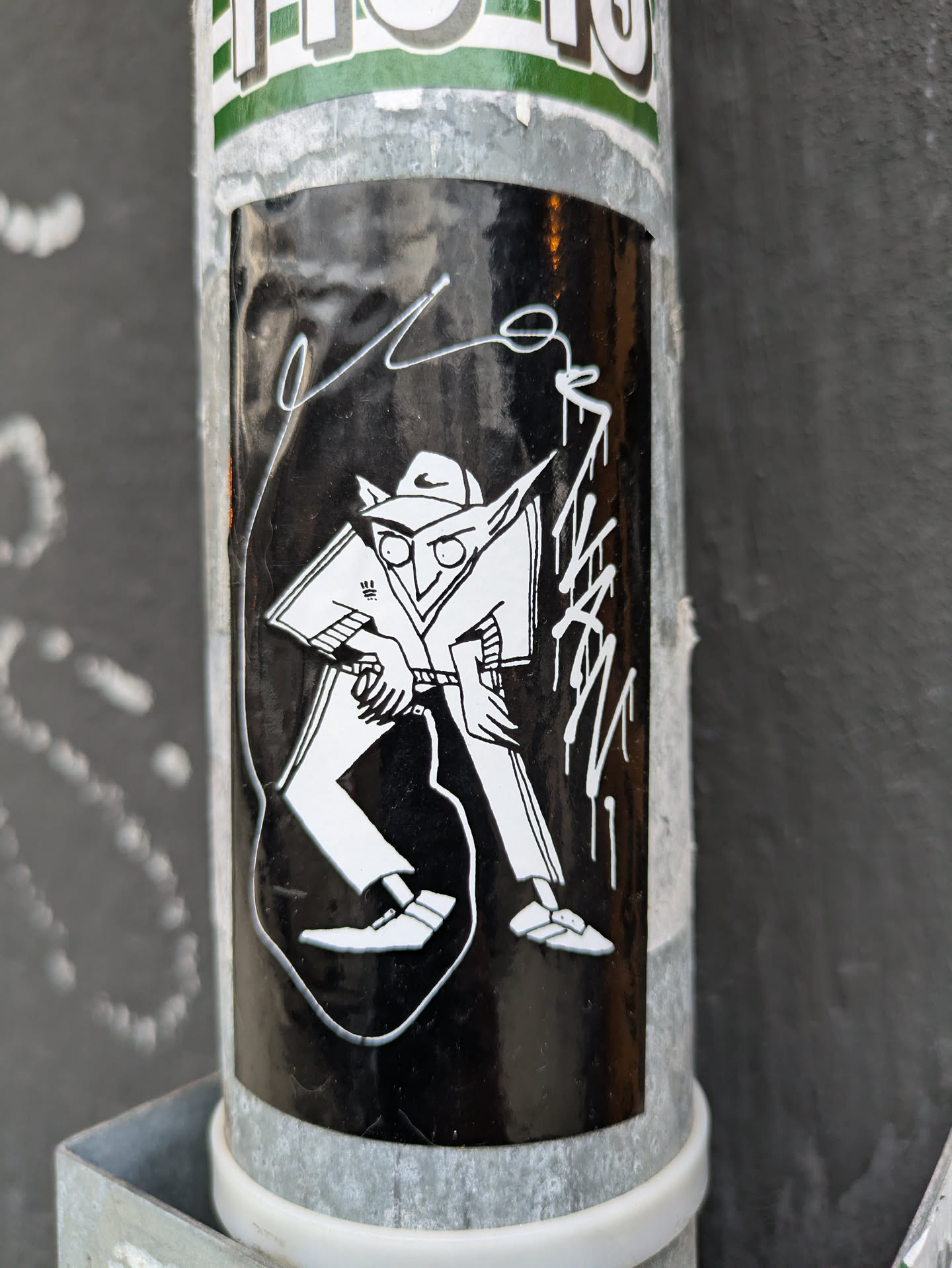 Black and white sticker featuring a stylized figure in a hat and loose-fitting clothing, possibly pants. The character's face is partially obscured, adding to the mysterious nature of the art. There are additional markings around the main figure, which could be considered part of the design or a signature. The sticker is affixed to a metallic pole.